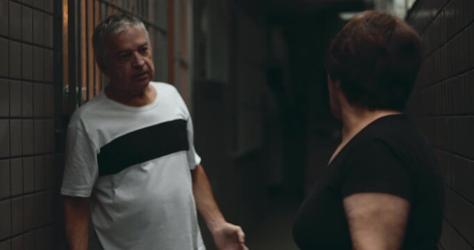 Elderly man dismissing argument with defensive body language, woman expressing anger with emphatic gestures, emotional conflict in tense hallway setting