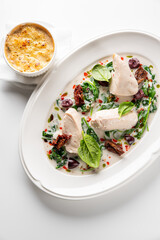 Creamy Tuscan chicken breast in a white sauce with spinach, sun-dried tomatoes, and olives on a white plate, with a side dish.