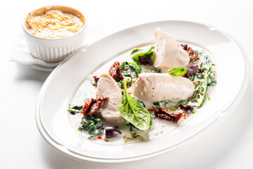 Creamy Tuscan chicken breast in a white sauce with spinach, sun-dried tomatoes, and olives on a white plate, with a side dish.