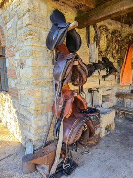 Rustic collection of worn, brown leather saddles and black tack hanging on a wooden rack against a rough, historic stone wall in a stable or rural tack room