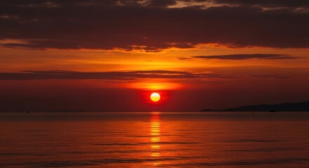 Obraz premium Fiery sun setting over a calm ocean reflects vibrant colors across the water.