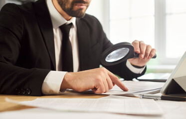 Business man auditor in office inspects documents with magnifier. He reviews paperwork and analysis...