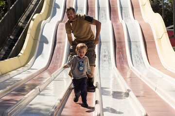 Father and son sliding down a playground slide on a sunny day, laughing and enjoying outdoor fun...
