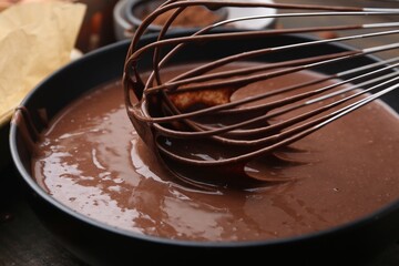 Mixing liquid chocolate dough with whisk in bowl at wooden table, closeup