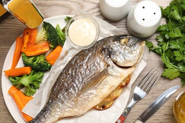 Tasty grilled fish served with vegetables and sauce on wooden table, flat lay