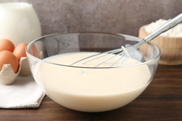 Liquid dough in bowl, whisk and ingredients on wooden table, closeup