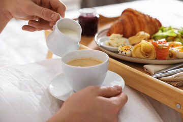Man pouring cream into coffee while having breakfast in bed, closeup. Hotel room service