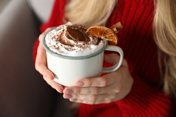 Beautiful woman having hot cocoa drink with whipped cream, cinnamon and orange slices in armchair at home, closeup