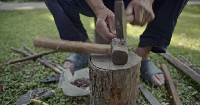 Captivating close-up footage of an older man diligently straightening old rusty nails on a wooden log in a grassy outdoor setting, accompanied by authentic hammering sounds.