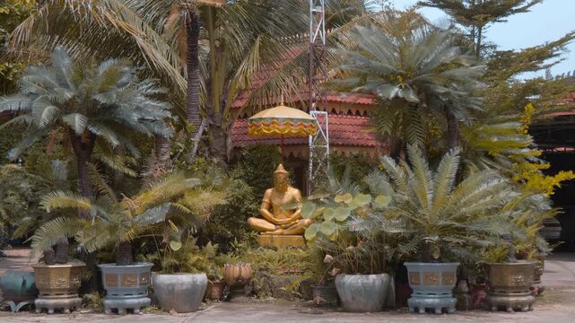 A gold colored statue of a meditating Khmer hermit sits under parasol in the temple garden of Wat Svay Romeat Pagoda in Siem Reap, Cambodia. Shot in motion