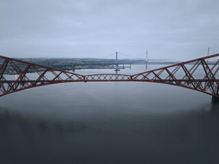 Aerial drone view of the Forth Bridge, Forth Road Bridge, and Queensferry Crossing over the Firth of Forth in Scotland. Iconic Scottish engineering landmarks on a cloudy day.