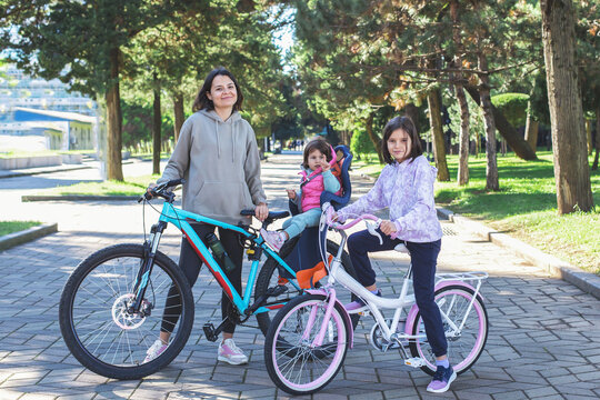Smiling woman with two children on bicycles in sunny park alley, one child sitting in bike seat. Family enjoying an active outdoor weekend together. Concept of healthy lifestyle, leisure, family time