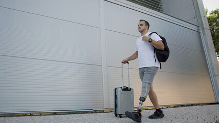 A young man walks confidently with a suitcase and backpack, showcasing his prosthetic leg, near a...