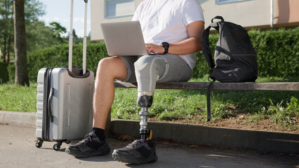 A person with a prosthetic leg sits on a bench using a laptop. A suitcase is beside them in a park...