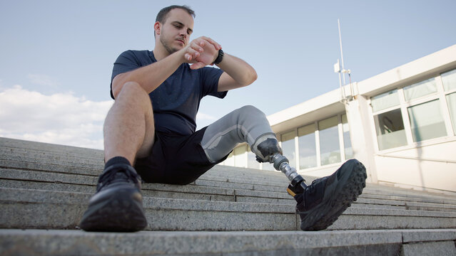 A man with a prosthetic leg sits on stairs, checking his watch while enjoying a sunny day outdoors in a modern area.