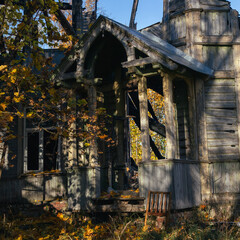 Front view of an old abandoned wooden house with a gothic-style porch. The structure is weathered and partially collapsed, surrounded by autumn leaves and sunlight. 
