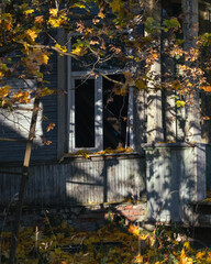 Old wooden house with broken window surrounded by colorful autumn leaves and sunlight casting shadows on the weathered walls.