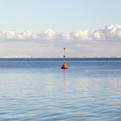 A bright red buoy floats in calm blue water under a soft cloudy sky, with a distant industrial coastline visible on the horizon.
