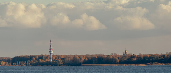 A scenic view of a television tower and a distant church on a forested shoreline, seen across a calm lake under cloudy skies.