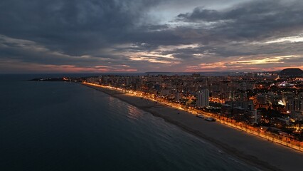 Beautiful aerial drone photograph showing a coastal city at dusk. The lights of the buildings and promenade reflect along the shoreline of the calm sea, 