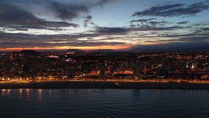 Beautiful aerial drone photograph showing a coastal city at dusk. The lights of the buildings and promenade reflect along the shoreline of the calm sea, 