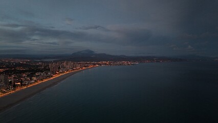 Beautiful aerial drone photograph showing a coastal city at dusk. The lights of the buildings and promenade reflect along the shoreline of the calm sea, 