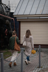 Two women walk near an old steam locomotive at the locomotive museum Povorotny Krug, one holding autumn leaves