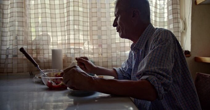 Captures an elderly man enjoying his meal alone at a kitchen table. The video includes natural sounds, enhancing the authenticity of the moment as he eats in the morning light.