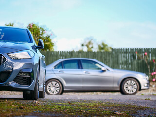 Two cars parked in a back yard of a country house. Cost of living out of the city in rural area. Expense of running two cars in a family theme. Need for transportation.