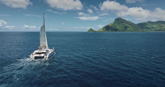 Fiji, Kuata island: White catamaran with tourists sailing blue ocean water near tropical island, under bright sunny sky. Remote wild nature paradise, exotic summer travel. Aerial drone flight footage