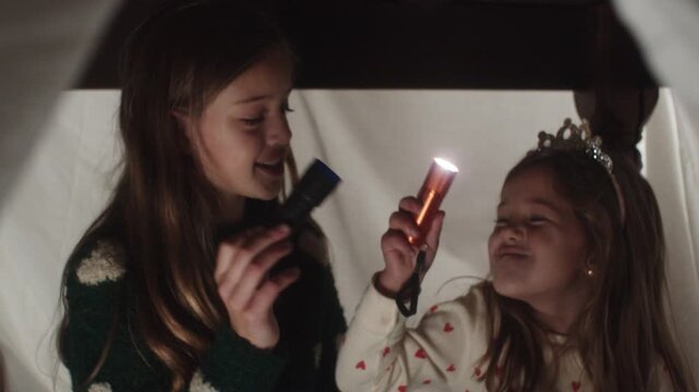Two girls enjoying flashlight fun under bedsheets at home in the evening