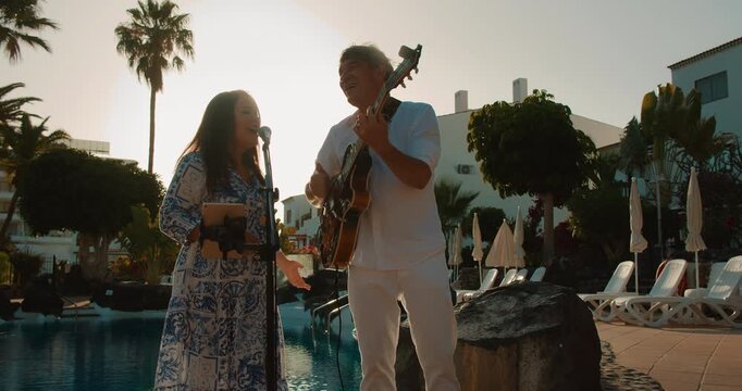 Man and woman perform music by poolside with backlight from evening sun