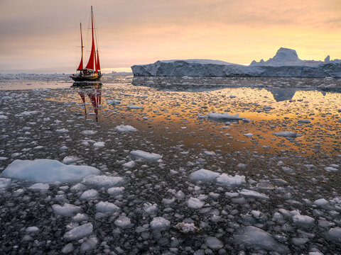 Red-sailed boat sailing through icy arctic waters surrounded by floating ice and large icebergs under cloudy sky.