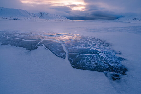 Frozen Ice Cracked and Covered with Snow in Foreground with Arctic Mountains and Dramatic Sky at Twilight, Svalbard