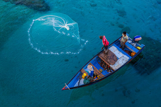 Two fishermen on colorful wooden boat casting net into crystal-clear sea from above