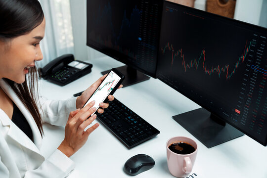 Smiling young Asian businesswoman with happy face looking on smartphone in profitable of exchange stock market graph at modern office. Concept of investing high profit in wealth security. Stratagem.