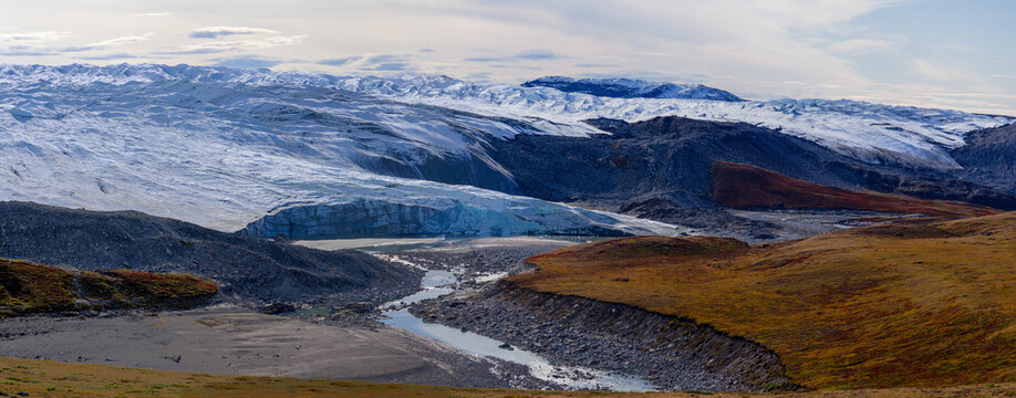 Panoramic view of glacier flowing into river valley surrounded by tundra under cloudy sky