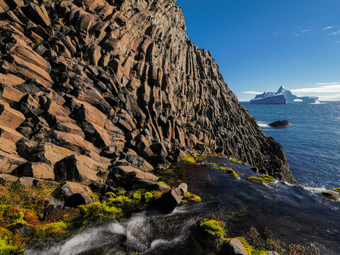 Dramatic basalt cliffs with moss and small stream overlooking iceberg in clear blue sea