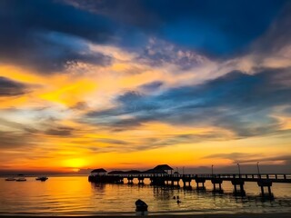 Breathtaking tropical sunset illuminates a serene pier stretching into calm waters, adorned with vivid orange, yellow, and blue skies, creating a peaceful evening landscape scene