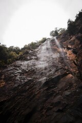 Powerful natural waterfall plunges dramatically down a dark, rugged cliff face, showcasing the raw beauty of untamed wilderness with hints of lush green foliage under an expansive sky