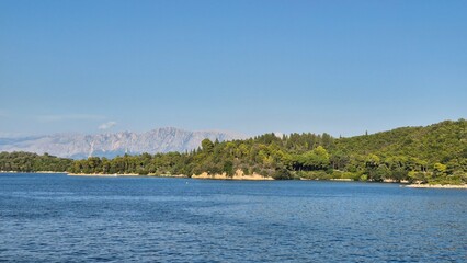 Stunning Mediterranean seascape with clear blue water, lush green forested coastline, and a backdrop of rugged mountains under a clear sky, suggesting a tranquil Greek island.
