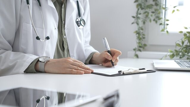 Professional female doctor wearing white lab coat and stethoscope documenting patient information on clipboard near laptop in clinic workspace. Medicine and health care concept - Powered by Adobe