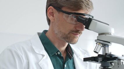 Senior man scientist wearing protective glasses and lab coat analyzing samples with a microscope in laboratory setting. Medicine, health care and science concept