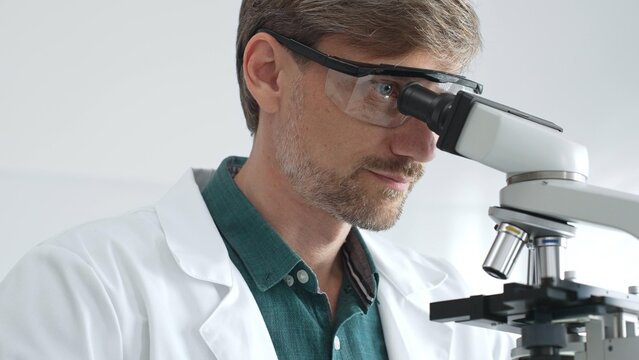 Senior man scientist wearing protective glasses and lab coat analyzing samples with a microscope in laboratory setting. Medicine, health care and science concept - Powered by Adobe