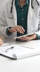 Male doctor in lab coat over a green shirt, and stethoscope gesturing while explaining diagnosis to patient using tablet in modern medical office. Medicine and health care concept