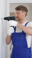 Professional male construction worker in a blue jumpsuit, safety goggles, and work gloves is screwing a screw into the wall using a cordless screwdriver, carrying out a careful home renovation
