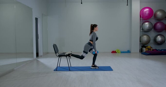 athletic woman is engaged in a Bulgarian split squat using dumbbells in an indoor gym. She focuses on her form while balancing on one leg with fitness equipment visible in the background.