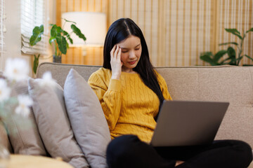 Upset brunette young asian woman sitting on couch at home, holding laptop and touching her head, looking at copy space, waiting for call from boyfriend or business partner, experiencing difficulties
