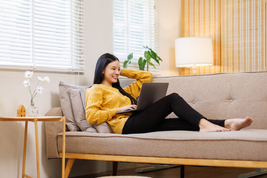 Photo of pretty young asian woman sit couch video call or work on laptop wear yellow pullover loft interior apartment indoors