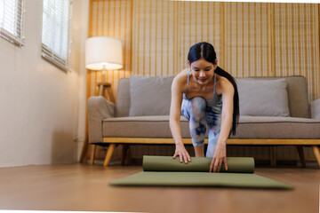 Female asian adult rolls yoga mat on wooden floor, suggesting preparation for exercise. Neutral...
